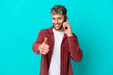 Genç, beyaz, mavi arka planda izole edilmiş genç bir adam. Cep telefonuyla konuşurken baş parmağını kaldırıyor.