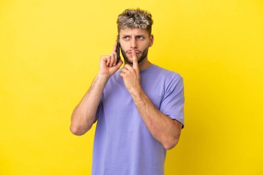 Sarı arka planda izole edilmiş cep telefonu kullanan genç beyaz adam yukarı bakarken şüpheleri var.