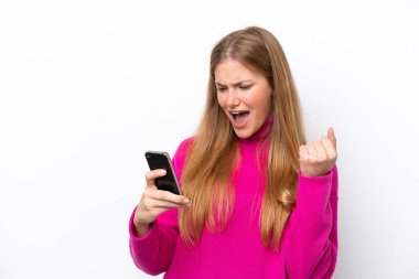 Beyaz arka planda izole edilmiş genç beyaz bir kadın ve telefon zafer pozisyonunda.