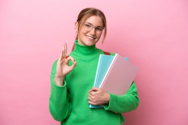 Pembe arka planda izole edilmiş genç beyaz kadın parmaklarıyla tamam işareti gösteriyor.