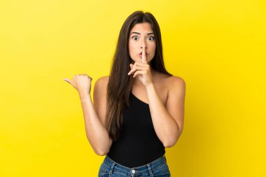Sarı arka planda izole edilmiş genç Brezilyalı bir kadın. Yan tarafı işaret ediyor ve sessizlik hareketi yapıyor.