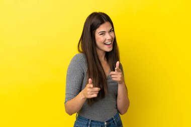 Sarı arka planda izole edilmiş genç Brezilyalı kadın ön tarafı işaret ediyor ve gülümsüyor.