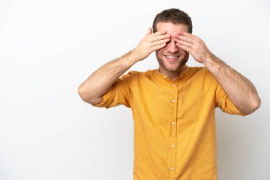 Beyaz arka planda izole edilmiş beyaz tenli genç bir adam. Gözleri elleriyle kaplıyor ve gülümsüyor.