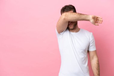 Pembe arka planda izole edilmiş genç, beyaz bir adam gözlerini elleriyle kapatıyor.