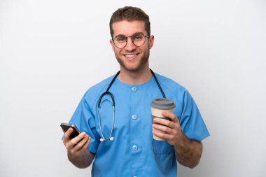 Genç cerrah, beyaz arka planda izole edilmiş beyaz bir adam elinde kahve ve cep telefonu var.