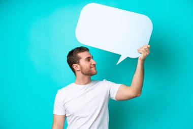 Mavi arka planda izole genç beyaz adam boş bir konuşma balonu tutuyor.