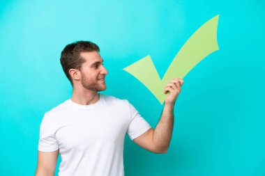 Mavi arka planda izole edilmiş genç beyaz bir adam elinde mutlu bir ifadeyle bir onay simgesi tutuyor.