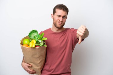 Beyaz arka planda izole edilmiş bir alışveriş poşeti tutan genç beyaz bir adam olumsuz bir ifadeyle baş parmağını gösteriyor.