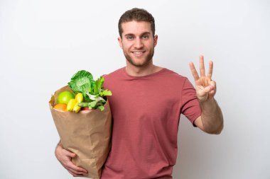 Beyaz arka planda izole edilmiş alışveriş torbası tutan beyaz bir adam mutlu ve parmaklarıyla üçe kadar sayıyor.
