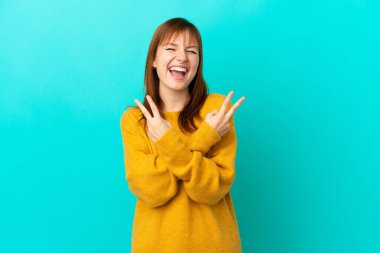 Mavi arka planda izole edilmiş kızıl saçlı kız gülümsüyor ve zafer işareti gösteriyor.