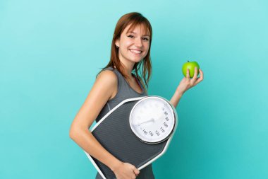 Kızıl saçlı kız mavi arka planda tartı makinesi ve elmayla izole edilmiş.