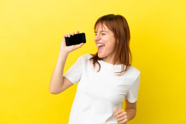 Kızıl saçlı kız sarı arka planda cep telefonu ve şarkı söyleyerek izole edilmiş.