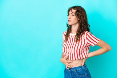 Kıvırcık saçlı genç bir kadın, mavi arka planda izole edilmiş, gayret gösterdiği için sırt ağrısı çekiyor.
