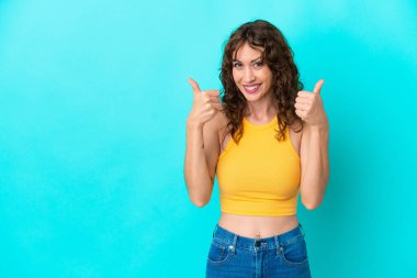 Mavi arka planda, kıvırcık saçlı genç bir kadın baş parmak hareketi yapıyor.