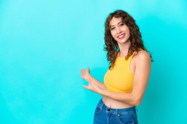 Mavi arka planda yalıtılmış kıvırcık saçlı genç bir kadın davet edilmek için ellerini yana uzatıyor.