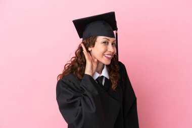 Üniversite mezunu genç bir kadın pembe arka planda izole edilmiş bir şekilde bir şeyler dinlerken elini kulağına koyar.