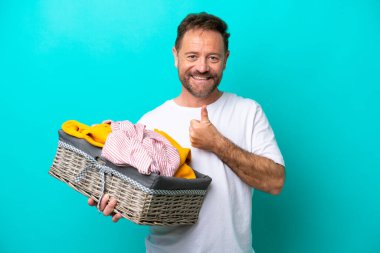 Mavi arka planda izole edilmiş çamaşır sepetini tutan orta yaşlı bir kadın baş parmak işareti yapıyor.