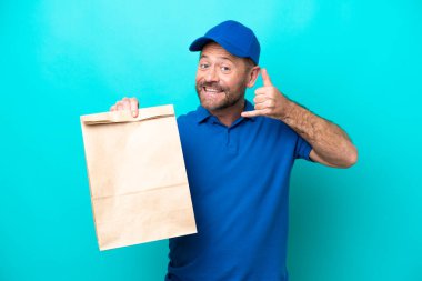 Orta yaşlı bir adam mavi arka planda izole edilmiş bir paket yiyecek alıyor ve telefon jesti yapıyor. Bana geri dönüş işareti ver.