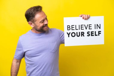 Middle age caucasian man isolated on yellow background holding a placard with text Believe In Your Self clipart