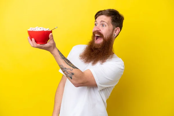 Redhead man with beard eating a bowl of cereals isolated on yellow background with surprise and shocked facial expression
