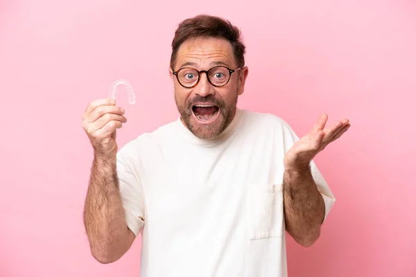 Middle age man holding invisible braces isolated on pink background with shocked facial expression