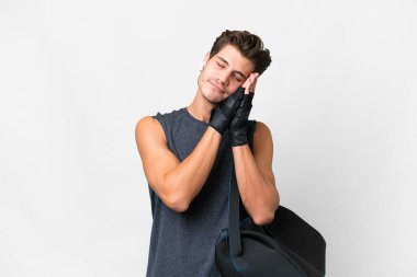 İzole edilmiş beyaz arka planda spor çantasıyla genç beyaz bir adam uyku jesti yapıyor.