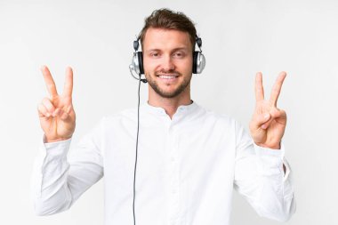Tele pazarlamacı beyaz kulaklıkla çalışan beyaz bir adam zafer işaretini iki eliyle de gösteriyor.
