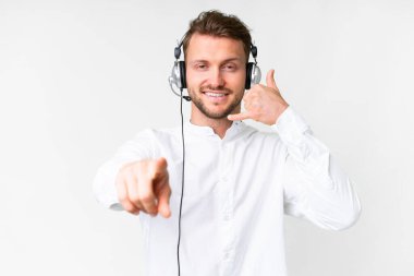 Tele pazarlamacı beyaz bir arka planda kulaklıkla çalışan beyaz bir adam telefon jesti yapıyor ve ön tarafı işaret ediyor.