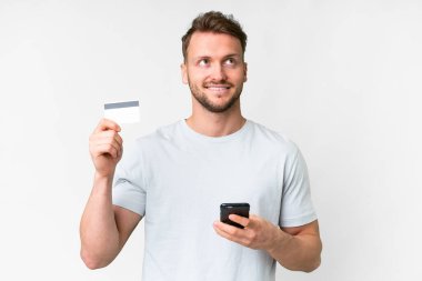 Beyaz arka planda yaşayan yakışıklı beyaz bir adam kredi kartıyla cep telefonundan alışveriş yapıyor.