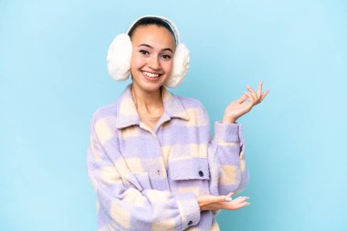 Mavi arka planda izole edilmiş kışlık kuklalar takan genç bir Arap kadını davet etmek için ellerini yana uzatıyor.