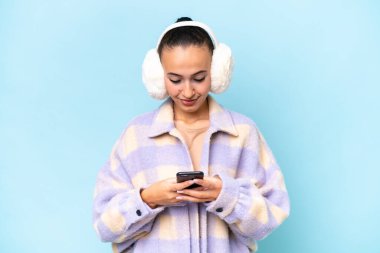 Mavi arka planda izole edilmiş kışlık manşet giyen genç bir Arap kadın cep telefonuyla mesaj gönderiyor.