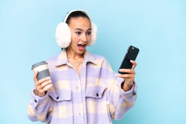 Mavi arka planda izole edilmiş kışlık kuklalar takan genç bir Arap kadın elinde kahve ve bir cep telefonu tutuyor.