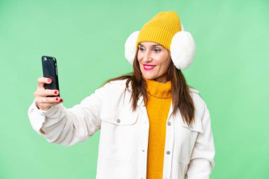 Orta yaşlı bir kadın, izole edilmiş krom anahtar arkaplan üzerine kışlık manşet takıyor ve selfie çekiyor.