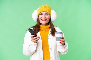 Orta yaşlı bir kadın, izole edilmiş krom arka planda kış maskesi takıyor. Elinde kahve ve cep telefonu var.