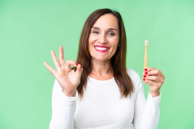 Dişlerini fırçalayan orta yaşlı bir kadın izole edilmiş krom anahtar arkaplanda parmaklarıyla tamam işareti gösteriyor.