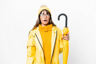 Yağmura dayanıklı ceketi ve şemsiyesi olan beyaz arka planda genç beyaz bir kadın yukarı bakıyor ve şaşırmış bir ifadeyle
