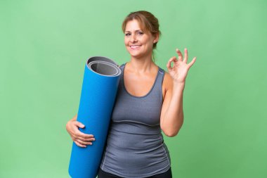 Orta yaşlı sporcu kadın yoga derslerine giderken arka planda izole edilmiş bir paspas tutuyor ve parmaklarıyla işaret veriyor.