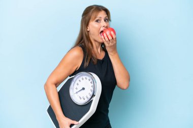 Orta yaşlı beyaz kadın, mavi arka planda, tartı makinesi ve elmayla izole edilmiş.