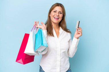 Orta yaşlı beyaz bir kadın mavi arka planda izole edilmiş. Elinde alışveriş çantaları ve bir kredi kartı var.