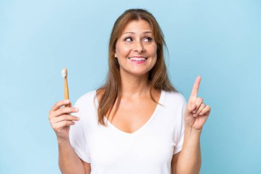 Mavi arka planda dişlerini fırçalayan orta yaşlı beyaz kadın çözümü bulmak için parmağını kaldırıyor.