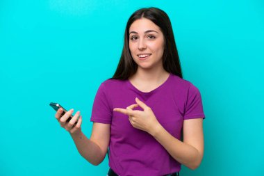 Young girl isolated on blue background using mobile phone and pointing it
