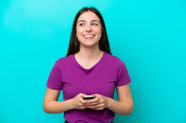 Young girl isolated on blue background using mobile phone and looking up