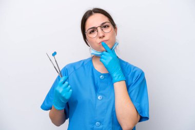 Beyaz arka planda izole edilmiş aletleri tutan beyaz dişçi kadın yukarı bakarken şüphe duyuyor