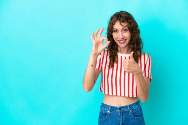 Mavi arka planda izole edilmiş kıvırcık saçlı genç kadın işaret ve baş parmak hareketi gösteriyor.