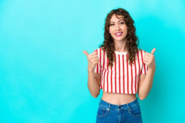 Kıvırcık saçlı genç bir kadın mavi arka planda izole edilmiş. Baş parmakları havada ve gülümsüyor.