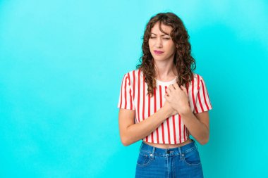 Mavi arka planda izole edilmiş kıvırcık saçlı genç bir kadın. Kalbinde ağrı var.