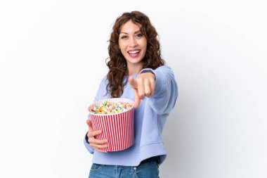 Beyaz arka planda izole edilmiş kıvırcık saçlı genç bir kadın elinde bir kova patlamış mısırla ön tarafı işaret ediyor.