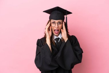 Üniversiteden mezun genç bir kadın pembe arka planda gözlüklü ve şaşırmış.