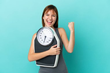 Kızıl saçlı kız mavi arka planda, tartı makinesiyle izole edilmiş ve zafer jesti yapıyor.