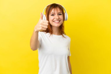 Kızıl saçlı kız sarı arka planda izole edilmiş müzik dinliyor ve başparmağını kaldırıyor.